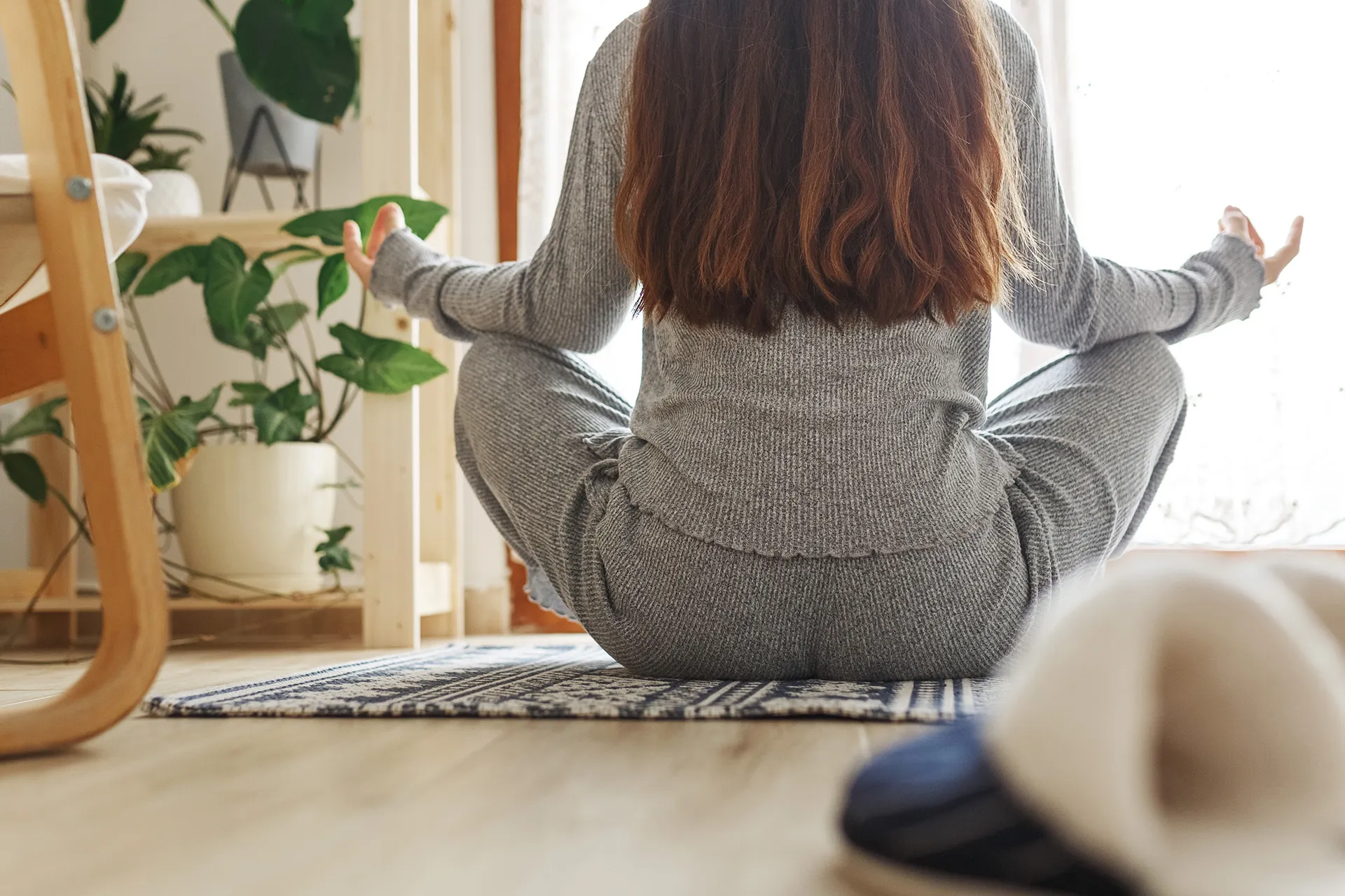 entspannte Person sitzt im Schneidersitz und meditiert im Wohnzimmer bei Tageslicht